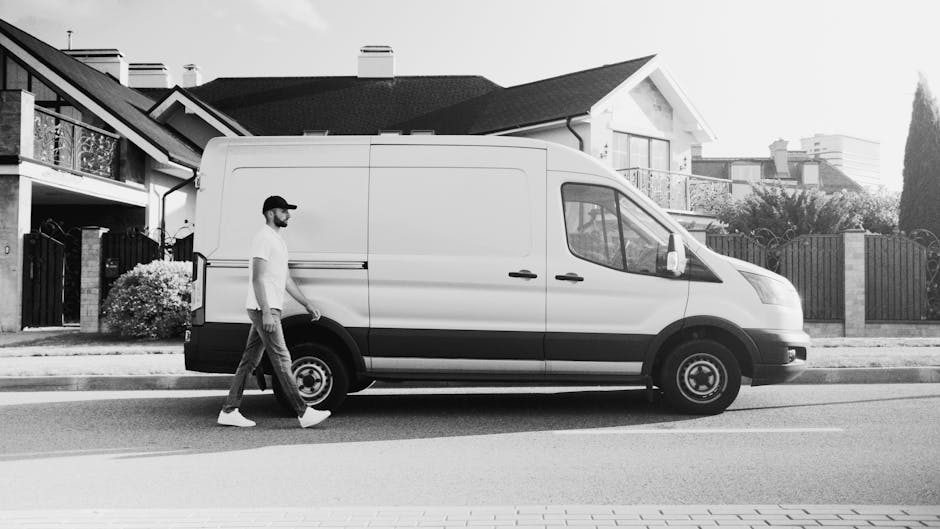 A white commercial van with a high roof, parked on a cobblestone driveway in a residential area during daytime. Inside the cab, a person wearing glasses and a dark jacket is seated, holding the steeri