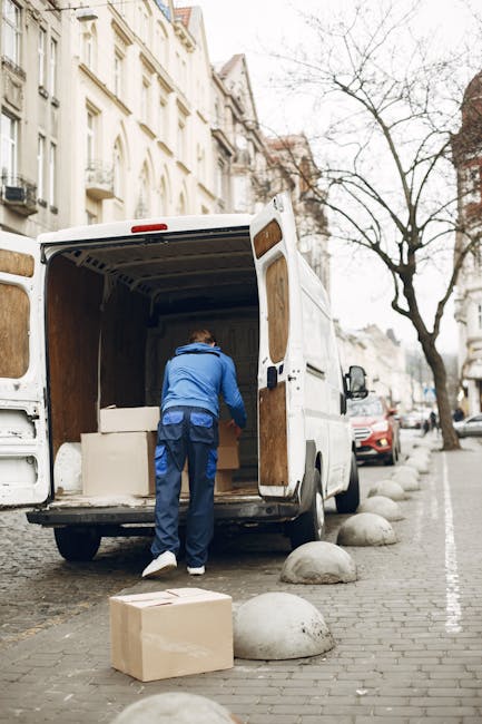 A man wearing a dark jacket is standing inside the open rear doors of a large white van, surrounded by numerous cardboard boxes of varying sizes that are stacked and arranged on the van's cargo area f
