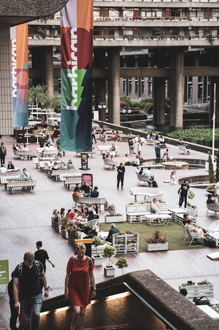 The image shows an outdoor urban area with multiple seating arrangements including benches and tables, all set upon a paved surface. Several people are seated, walking, or standing, engaging in variou