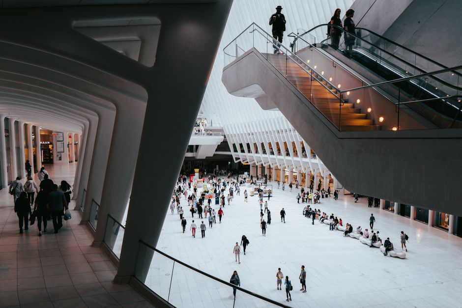 The image shows an outdoor urban area with multiple seating arrangements including benches and tables, all set upon a paved surface. Several people are seated, walking, or standing, engaging in variou