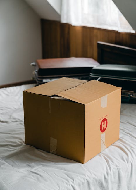 A person standing indoors during a home relocation process, shown lifting a medium-sized cardboard box with labeled sides onto a stack of similar boxes. The individual is wearing light-colored clothin