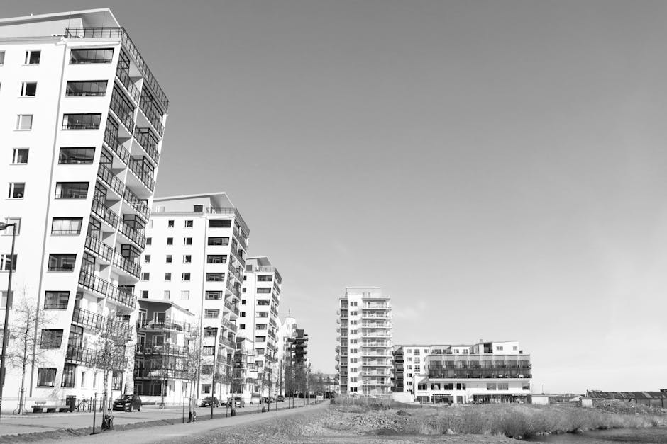 Black and white photograph depicting a residential area with multiple modern multi-storey apartment buildings lining the street. In the foreground, there are several open cardboard boxes, along with furniture items wrapped in protective fabric and plastic, positioned on a paved pavement area. A hand truck or trolley is visible, used for loading or transporting these items. The scene captures the home relocation process, with furniture and packing materials prepared for moving. A man from Man and Van White City can be seen loading or unloading furniture into or from a large van parked on the street, which is partially visible at the edge of the image. The environment is well-lit, with clear skies overhead, illustrating a typical moving day environment that includes details of the furniture transport and packing aspects associated with professional removals services.