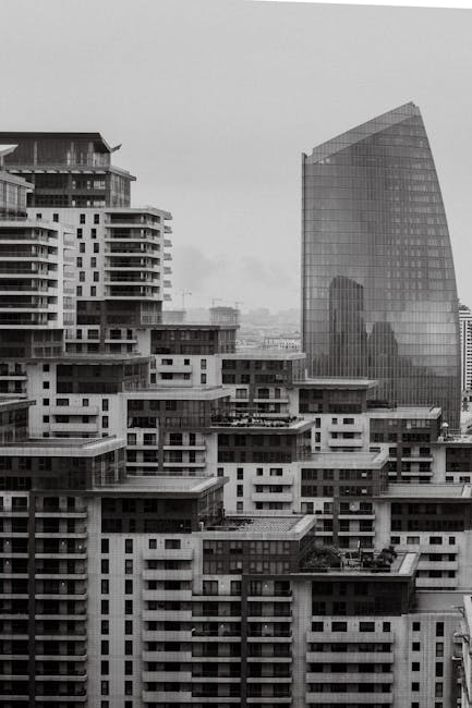 A black and white photograph of a cityscape featuring a collection of modern residential and commercial buildings. In the foreground, there are multiple mid-rise apartment blocks with flat rooftops, some with visible balconies and rooftop terraces. Behind these, a taller glass office or hotel building with a reflective surface that mirrors the surrounding structures and sky rises prominently. The scene appears to be taken from an elevated position, possibly from inside a property or near an upper floor, looking towards the urban skyline. The image suggests an environment associated with city living and urban development, relevant to house removals or home relocation services. Man and Van White City could utilize this visual to illustrate urban moving logistics, multi-property transfers, or the complexity of moving within a high-density area.