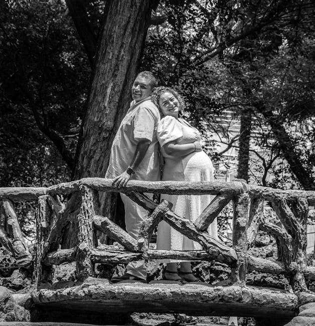 Black and white photograph depicting a residential area with multiple modern multi-storey apartment buildings lining the street. In the foreground, there are several open cardboard boxes, along with furniture items wrapped in protective fabric and plastic, positioned on a paved pavement area. A hand truck or trolley is visible, used for loading or transporting these items. The scene captures the home relocation process, with furniture and packing materials prepared for moving. A man from Man and Van White City can be seen loading or unloading furniture into or from a large van parked on the street, which is partially visible at the edge of the image. The environment is well-lit, with clear skies overhead, illustrating a typical moving day environment that includes details of the furniture transport and packing aspects associated with professional removals services.